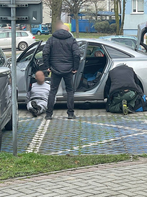 Zatrzymany w kajdankach leży oparty o samochód. Obok dwaj funkcjonariusze - jeden stoi drugi kuca.
