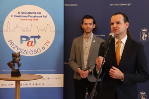 Na zdjęciu mężczyzna w trakcie przemowy. W tle stoi mężczyzna, obok niego stolik ze statuetką - symbolem programu.